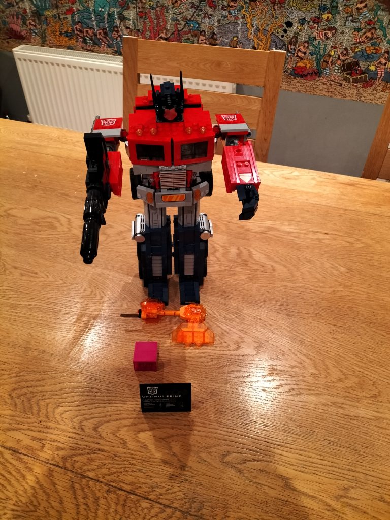 The completed model, standing in robot form, with gun in hand. Axe and Energon cube on the table in front of the robot, and a plaque about Optimus Prime.