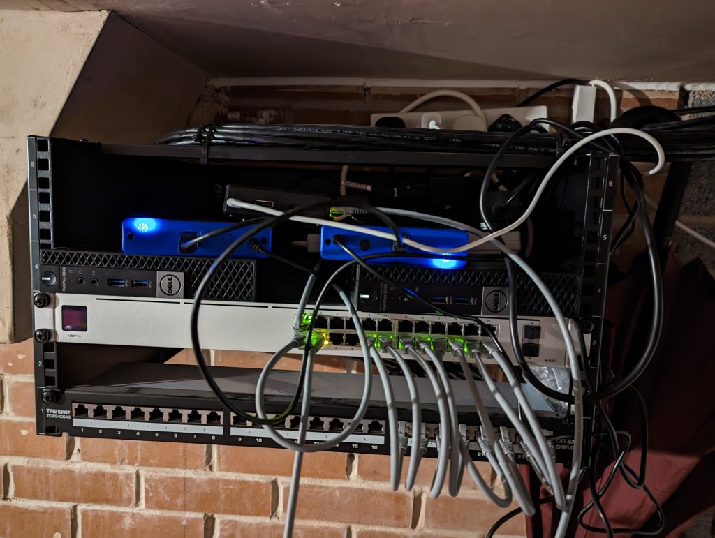 Picture of a comms rack with a patch panel, a unifi USW-Pro-24 switch, two Dell Optiplex 3040M computers, two external hard drives and a Raspberry Pi.
